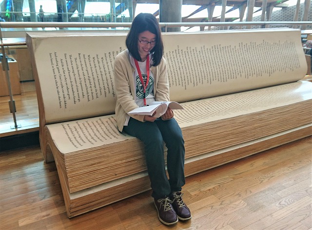 小澤彩果さんが本の形のベンチで読書している写真